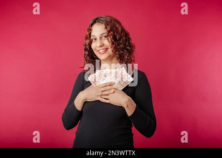 Portrait of young redhead woman wearing black dress standing isolated over red background holding banknotes near chest, laughs and looks at the camera Stock Photo