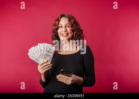 Happy woman wearing black dress standing isolated over red background showing bunch of money banknotes and holding mobile phone. Looking to the camera Stock Photo