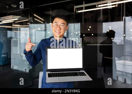 Portrait of smiling successful asian man inside office, man smiling and looking at camera standing holding laptop and pointing finger at white screen, businessman in shirt and glasses at workplace. Stock Photo