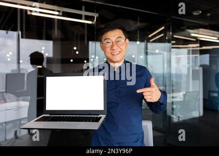 Portrait of smiling successful asian man inside office, man smiling and looking at camera standing holding laptop and pointing finger at white screen, businessman in shirt and glasses at workplace. Stock Photo