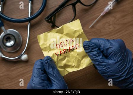 A hand wearing blue glove opening a crumpled yellow memo note written with Delta Variant. Stock Photo