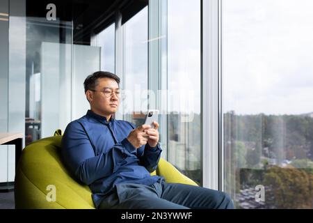 Serious thinking asian man reading news on phone, programmer using app on smartphone testing new program, businessman worker sitting on soft chair near window in modern office interior. Stock Photo