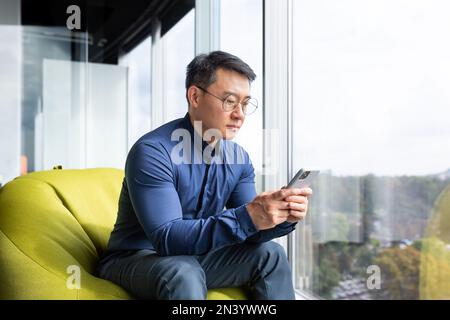 Serious thinking asian man reading news on phone, programmer using app on smartphone testing new program, businessman worker sitting on soft chair near window in modern office interior. Stock Photo
