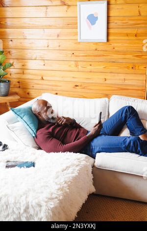 Smiling bald african american senior man using cellphone while relaxing on couch at log cabin Stock Photo