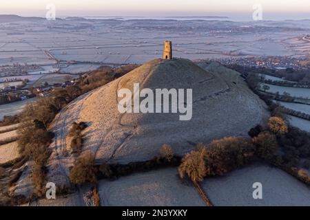 People climb the windy path up towards St Michael's Tower at the top of Glastonbury Tor as the sun rises over the Somerset Levels on a cold and crispy morning in many parts of the UK. Picture date: Wednesday February 8, 2023. Stock Photo