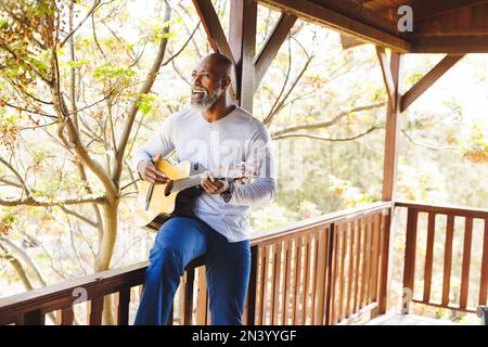 Bald african american senior man playing guitar and singing while sitting on railing in balcony Stock Photo
