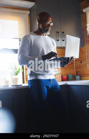 Serious african american bald senior man wearing eyeglasses using laptop while standing in kitchen Stock Photo