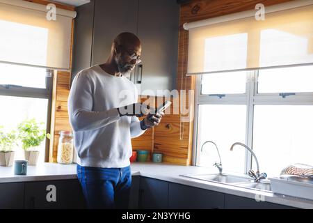 African american senior man wearing eyeglasses using mobile phone while standing by kitchen counter Stock Photo