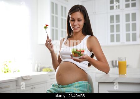 Young pregnant woman with bowl of vegetable salad at table in kitchen. Taking care of baby health Stock Photo