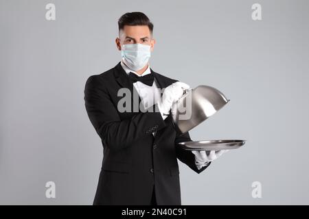 Waiter in medical face mask with empty tray on light grey background Stock Photo