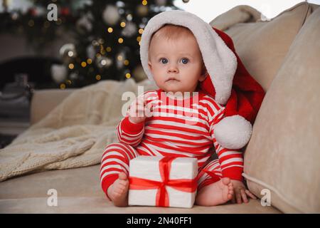 Cute baby in Santa hat with Christmas gift on sofa at home Stock Photo