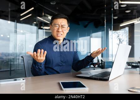 Portrait of frustrated and angry boss inside office, asian man looking at camera and shouting upset, man at workplace using laptop unhappy with work result. Stock Photo