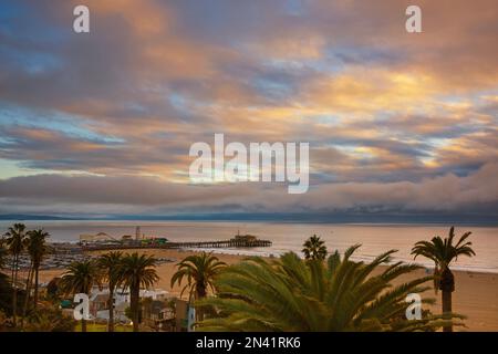 Low lying clouds kiss the ocean during a sunset that is illuminating a pier of amusement rides, sands of a beach and palm trees that line a road in th Stock Photo