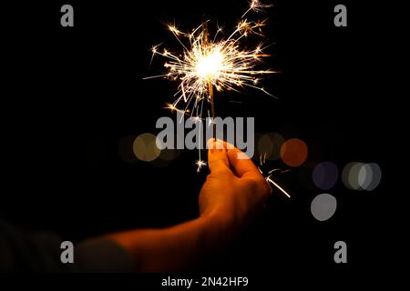 Woman holding bright burning sparklers against blurred lights, closeup Stock Photo
