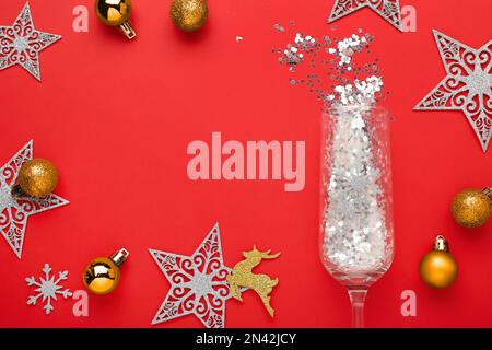 Flat lay composition with shiny confetti spilled out of champagne glass near Christmas decorations on red background, space for text Stock Photo