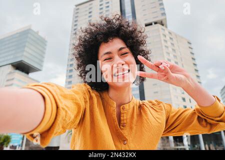 Young hispanic woman with curly hair, yellow shirt and cheerful attitude, smiling and having fun taking a selfie photo doing the peace sign with the fingers at city street outdoors. Lifestyle concept. High quality photo Stock Photo