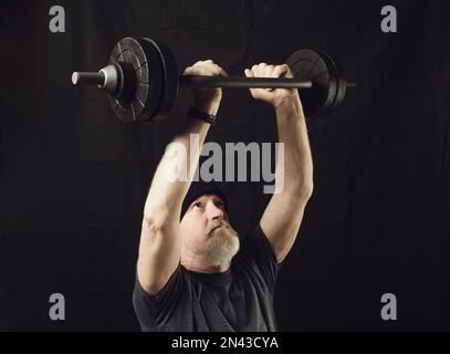 Fitness, an elderly bearded man holds a barbell in his arms straightened above his head. Stock Photo