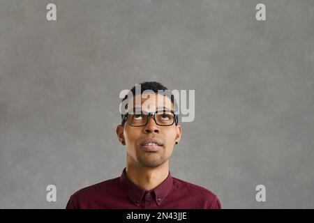 Dark-skinned man with doubtful facial expression looking up on gray concrete background. Stock Photo