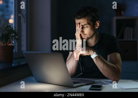 Young businessman has headache while working overtime with laptop at home office late night. Stressed depressed freelancer touching his head, feeling pain in eyes. Insomnia, internet addiction Stock Photo