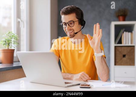 Guy wearing headset, start lesson, looking at laptop screen, waving hand, greeting tutor, has online class via video call, get skills through internet, remote education distantly with modern tech Stock Photo