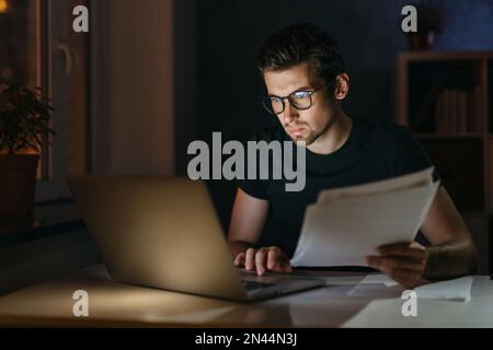 Portrait of tired focused young man in glasses working remotely late at night at home office with computer, looking at laptop, thinking about deadline. Overwork, negative thoughts, deadline concept Stock Photo