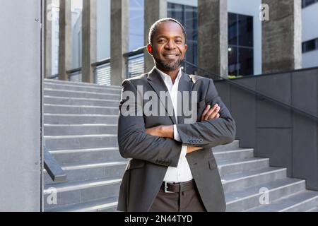 Portrait of a successful African American businessman. Standing in a suit outside the office center. He looks confidently into the camera, crosses his arms, smiles. Stock Photo
