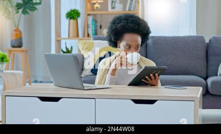 African American woman working remotely sitting on the floor having coffee with her tablet in her hand , laptop on coffee table and her back against Stock Photo