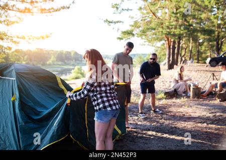 A company of friends guys and girls set up a tent on the sandy bank of the river in summer. Hiking, outdoor recreation in the forest. Sunny day, count Stock Photo