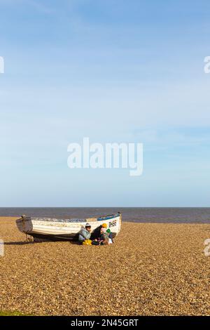 Single Disused Fishing Boat at Rest on Beach at Aldeburgh on a Bright and Sunny February Day with Young Family Sheltering from the Wind Stock Photo