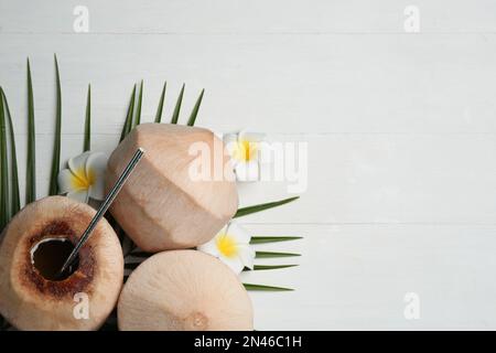 Young peeled coconuts with straw, tropical leaf and flowers on white wooden table, flat lay. Space for text Stock Photo