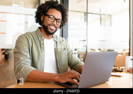 Portrait of smiling positive male office employee with computer indoors, African-American guy using laptop for remote work sitting in the modern coworking space, freelancer enjoys work on the distance Stock Photo