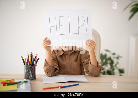 Bored upset tired caucasian little child sits at table shows paper with inscription help, suffer from overwork Stock Photo