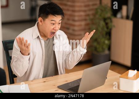 Great online offer. Emotional asian man using laptop and shouting, reading news, sitting at home and spreading hands Stock Photo