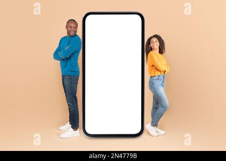 Black couple leaning on giant smartphone with blank screen, promoting mobile application or website, mockup Stock Photo