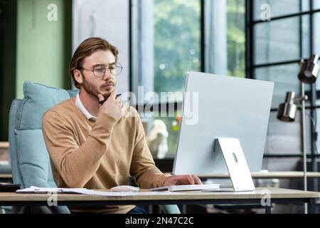 Portrait of a young designer, architect, engineer sitting in a modern office, working at a computer. He holds his chin with his hand, looks intently and thoughtfully at the monitor. Stock Photo