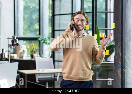 A successful young businessman, programmer, freelancer is standing in a modern office, talking on the phone, gesturing with his hands, smiling. Stock Photo