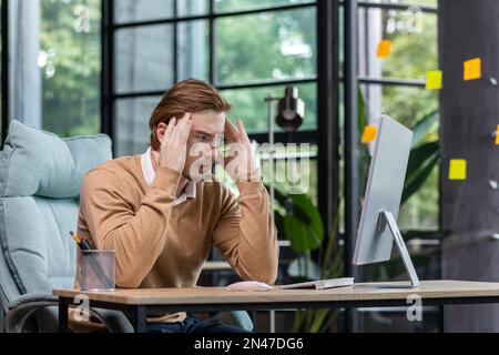 A young male programmer, IT developer codes, thinks about a new project. He is sitting concentrated at the desk in the office, holding his head thoughtfully with his hands, looking at the monitor. Stock Photo