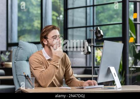 Successful young man, programmer, IT developer, software sitting at a desk in a modern office, working focused and thoughtful on a computer. Stock Photo