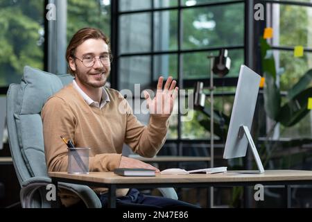 Portrait of a young businessman, designer, programmer in a busy office sitting at a table with a computer, smiling at the camera, waving, greeting, introducing himself. Stock Photo