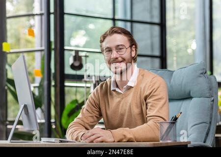 Portrait of a successful young man, programmer, IT developer, software sitting at a desk in a modern office, working at a computer. He looks at the camera, smiles. Stock Photo