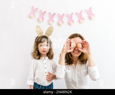 Mother and daughter with Easter decorations having fun with eggs. Cute bunny garland on the white background. Easter and Holidays concept Stock Photo