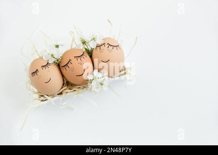 Brown hand drawn eggs with faces in the nest, with blooming flowers. Easter, Holiday concept, gift card for your design, place for text. White minimal Stock Photo
