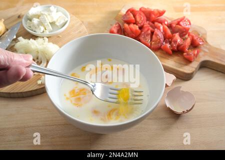 Eggs and milk are mixed with a fork in a white bowl on a wooden kitchen board, cooking preparation for an omelet or frittata with tomatoes and onions, Stock Photo