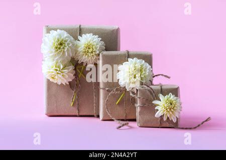 Three gift boxes in craft paper with white dahlia flowers on a gradient purple-pink background. The concept of a holiday, Valentine's day, women's day Stock Photo