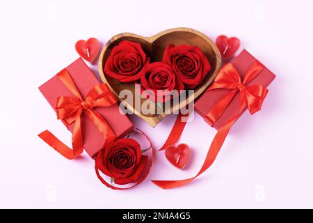 Three red roses in a wooden bowl in the shape of a heart, two red gift boxes with a red satin bow and candles in the shape of hearts on a pink backgro Stock Photo