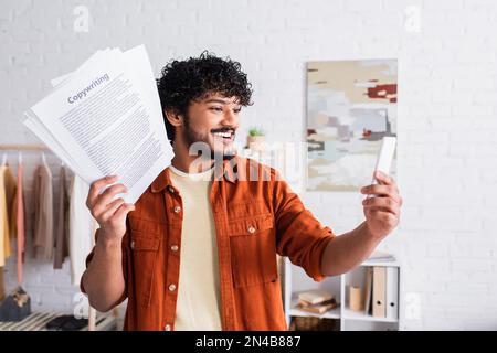 Smiling indian copywriter having video call on smartphone and holding papers at home,stock image Stock Photo