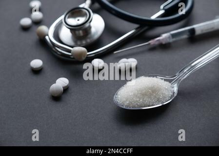 A white sugar in a transparent spoon together with stethoscope, pills and syringe on a black background. Stock Photo