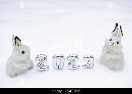 Two white cute and fluffy bunny toys with silver numbers 2023 on the white snow background. Copy space. Symbol of Chinese New Year 2023. Stock Photo