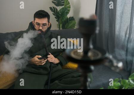 Bearded man is smoking hookah at home and blowing cloud of smoke - chill time and resting concept Stock Photo