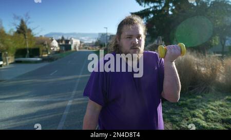 A young fat man getting back into shape building strength. Motivational concept of an overweight person listing weight dumbbells standing outside Stock Photo